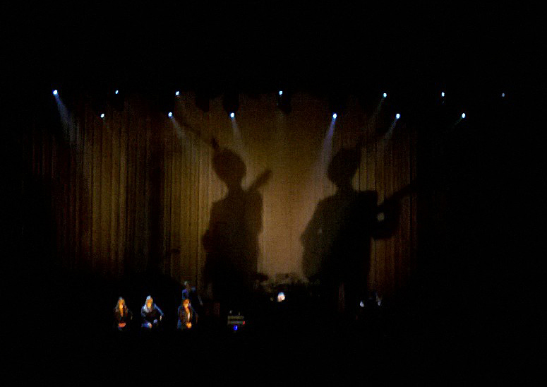 A spot of fancy lighting cast dual silhouettes of Leonard Cohen playing guitar.